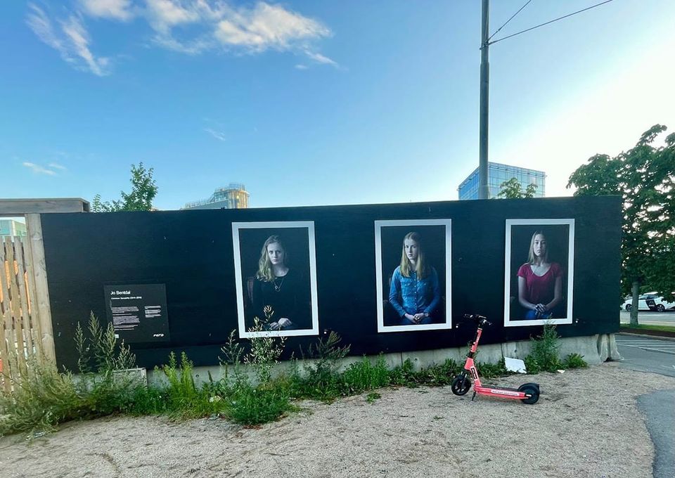 Portraits on a temporary wall outdoors, weeds growing before them and a pink scooter alongside, beautiful blue skies above
