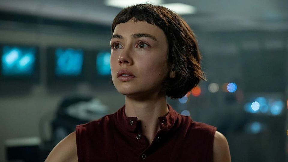 Soulful twenty-something lady with a short bob in a sleeveless top, staring outward, behind her blurry screens on a wall