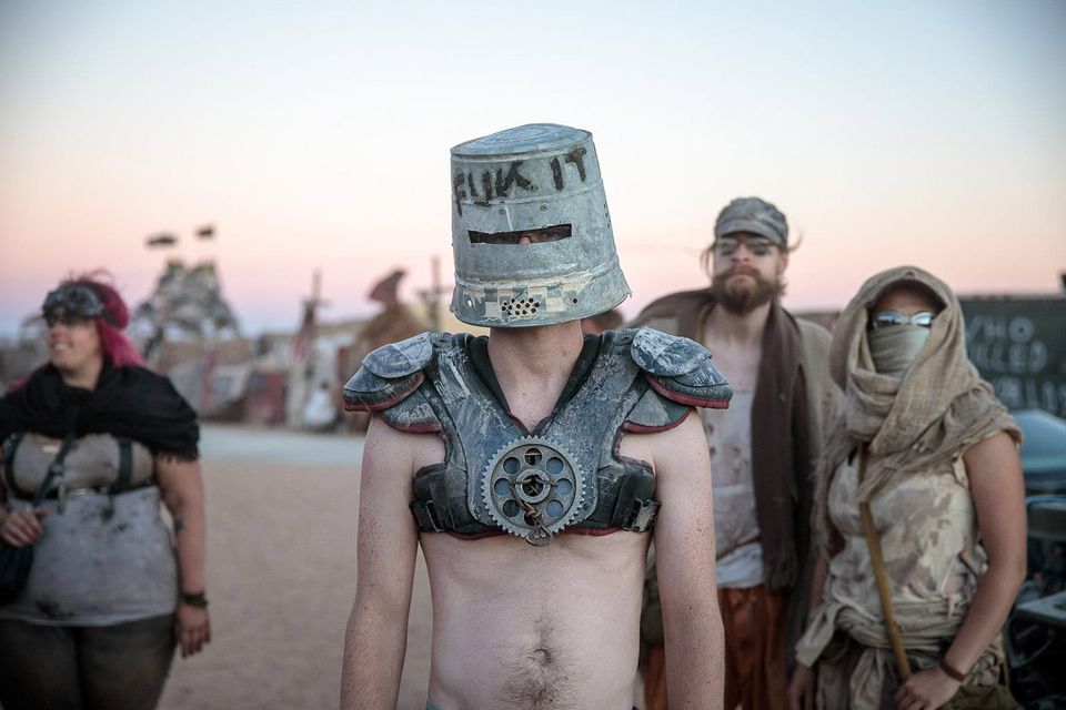 Mad Max Beyond Thunderdome desert-looking folks behind a person with a metal pail on their head with eye slit and FUK IT scrawled on front