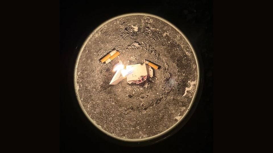 Cigarette butts and some burning papers in a sandy ashtray surrounded by darkness