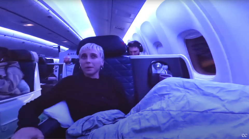 The influencer in a blonde pixie cut and long-sleeved pullover, reclining nervously under a throw in a dark blue-lit first class airplane seat