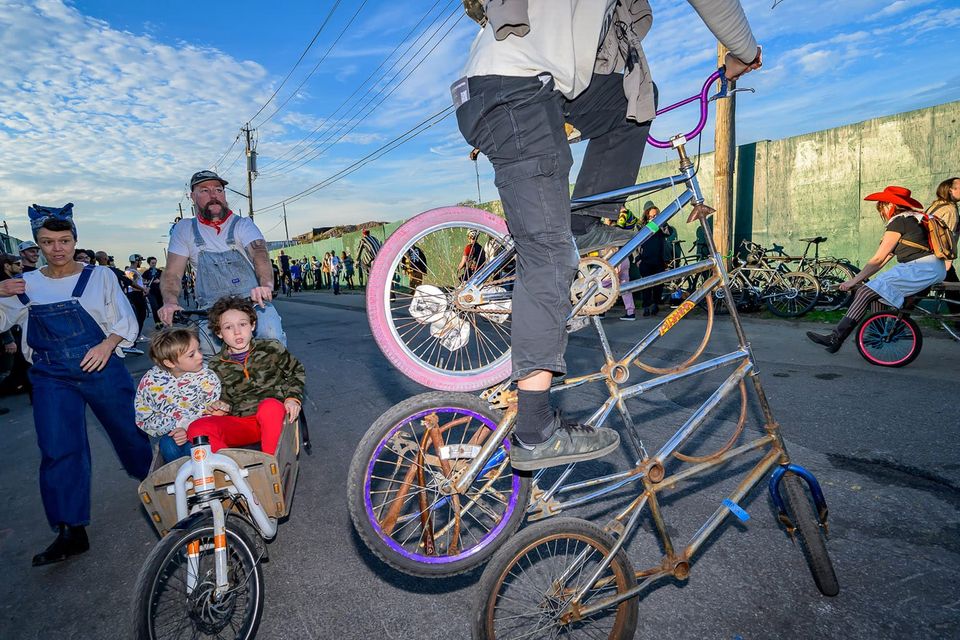 Triple high bicycle ridden by daring participant in Bike Kill, with other riders in the background against an enamel blue sky