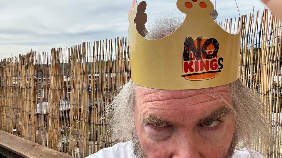 A man, outdoors, with greying hair and luxuriant eyebrows, wearing a NO KINGS hat (very cleverly modified from a Burger King cardboard hat) and a fierce expression