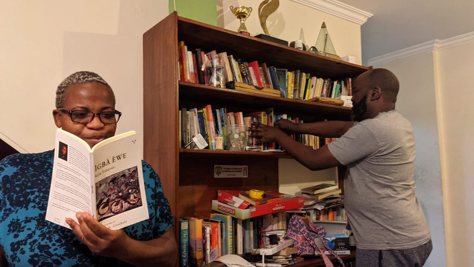Zukiswa Wanner in a blue dress, reading Kọ́lá Túbọ̀sún's 2021 poetry collection, Ìgbà Èwe. Behind her, a man in a grey T-shirt stands before a full bookshelf