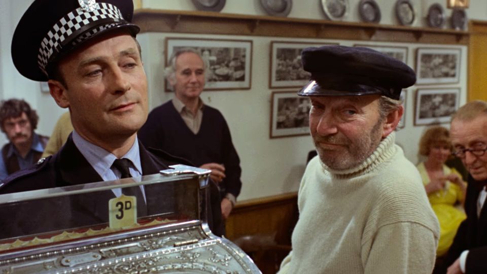 Edward Woodward as Sgt Howie in THE WICKER MAN (1973), in a pub full of locals, a cash register in the foreground and a grizzled, skeptical fisherman beside him