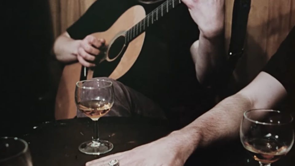 Screenshot from THE WICKER MAN (1973) shows a detail from the pub scene, with half-filled wineglasses, a guitar being strummed, a slender hand with a ring on it on a pub table