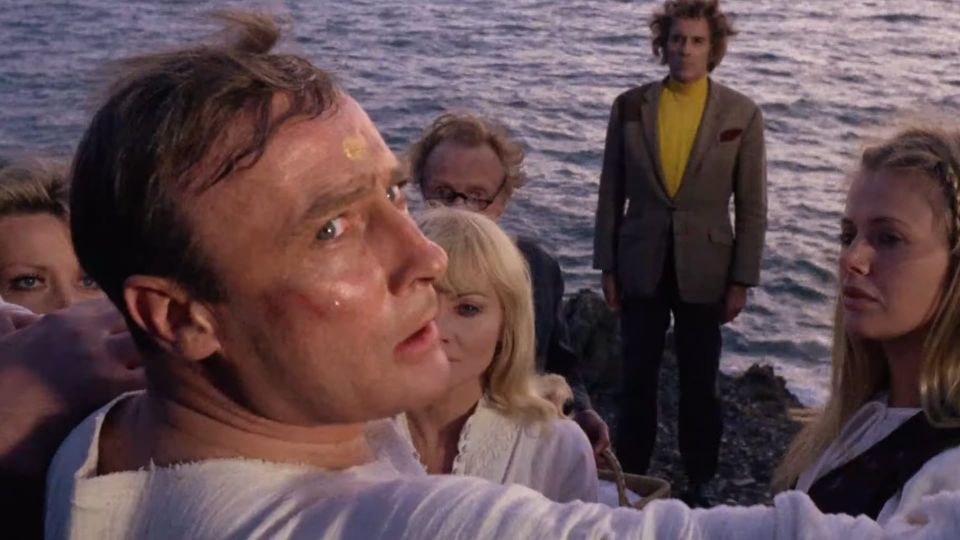 Screenshot from THE WICKER MAN shows Edward Woodward, face bruised and forehead stained with a ritual mark, wearing a white shift and an intent expression, the darkening sea behind him