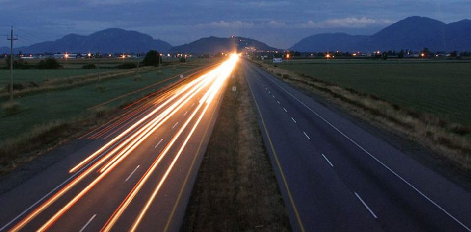 Streaks of car headlights in the eastbound lane, no cars in the westbound lane, sky beginning to lighten and reveal green fields on either side of the highway and mountains in distance.