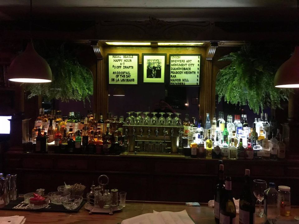 A dark bar, framed signs with specials and beer list, bottles and glasses flanked by ferns. REGAL BEAGLE HAPPY HOUR 4-7 $2 OFF DRAFTS - $8 COCKTAIL OF THE DAY - DE LA LOUISIANE