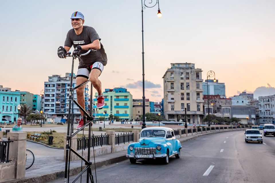 Félix Ramón Guirola Cepero riding a tall bike along a Havana highway near a small public park. Colorfully-painted multi-story buildings and an old car from the 40s or 50s in the background.