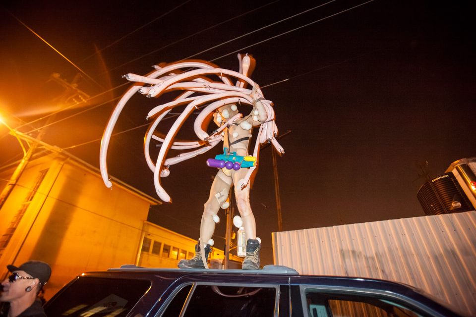 Reveler in a mad riotous ensemble of pink snake balloons and a super soaker standing on a car