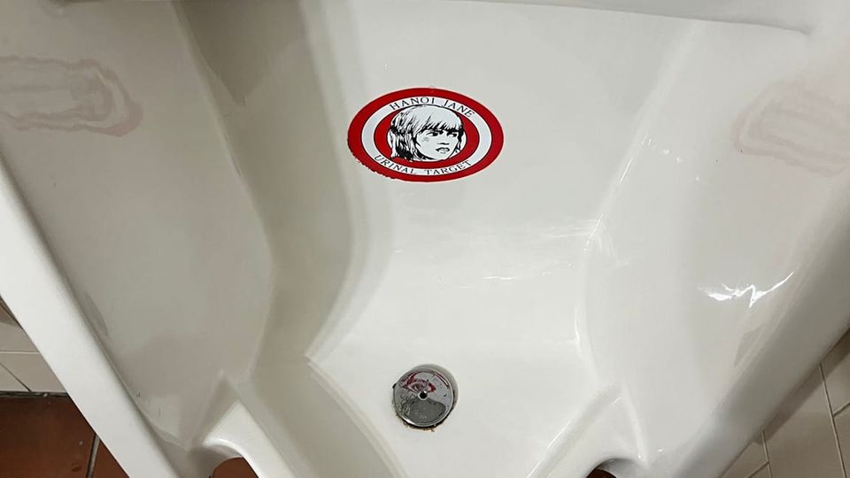 Photo of round HANOI JANE URINAL TARGET sticker complete with drawing of Jane Fonda inside concentric red circles affixed inside a urinal. 