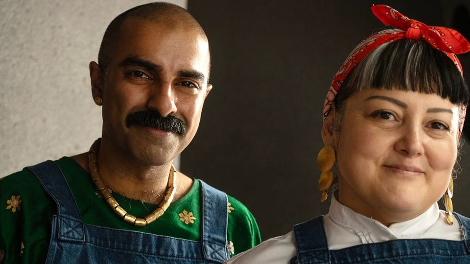 A smiling couple: man with a lush moustache and shaved head, in apron and patterned shirt; woman with bangs, in apron and red kerchief
