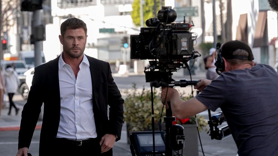 Chris Hemsworth character Davis walking in an exterior daylight street scene and we can see the cameraman filming.