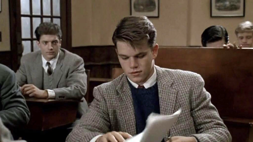 Students seated in class wearing oxford shirts neckties and tweed jackets. Matt Damon in foreground looking at papers, Brendan Fraser farther back looking at him.