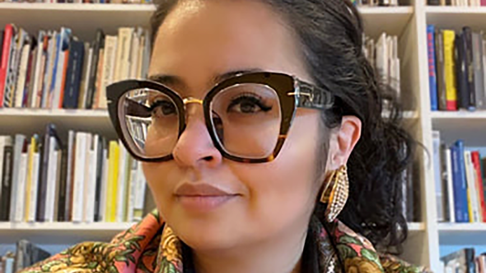 A lady in ginormous glasses, big earrings, richly patterned colorful scarf, lovely smile, posting before a book-lined wall