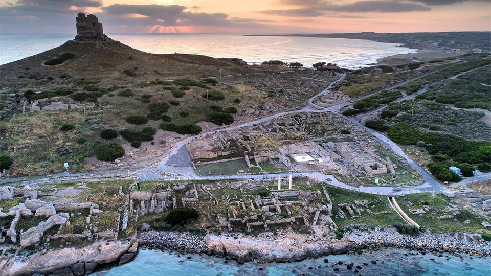 A vast excavation on the island of Sardinia shows evidence of a complex civilization with the sea and a medieval castle visible in the background