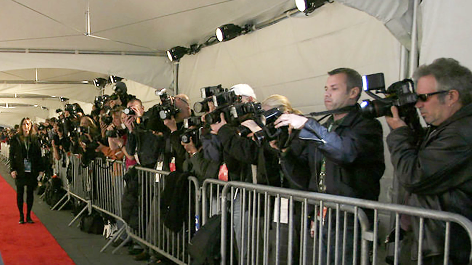 Paparazzi clicking away lined up along a metal barricade next to a red carpet at a film festival premiere