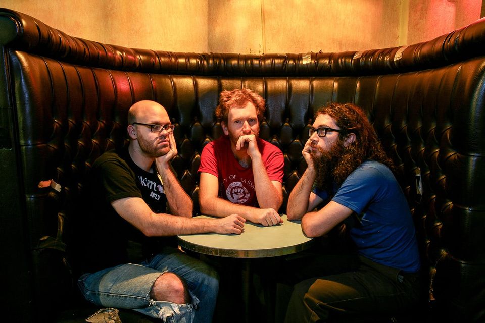 Three band members sitting in a semi-circular banquette looking unimpressed with everything