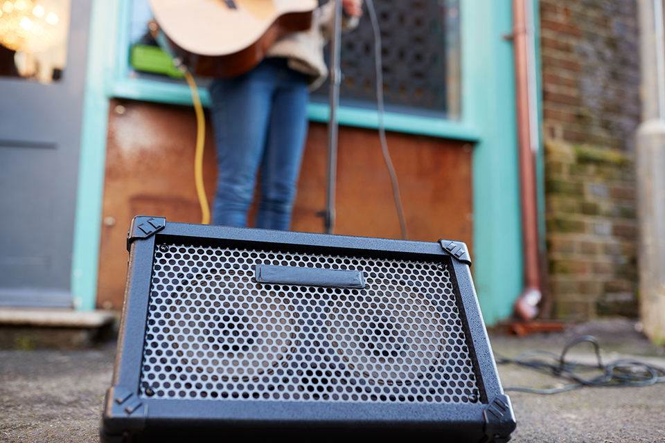 A loudspeaker in the street, with the legs and guitar of a street musician visible behind