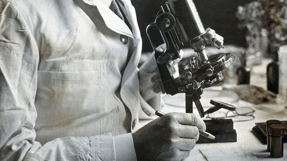 Detail of a 1930 photograph of a scientist in a white coat seated before a microscope