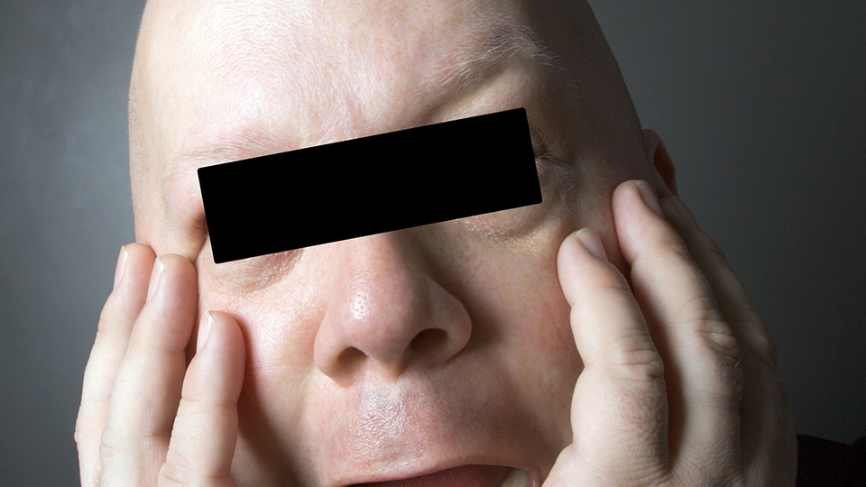 Distraught head-shaved individual with black bar over eyes to protect identity