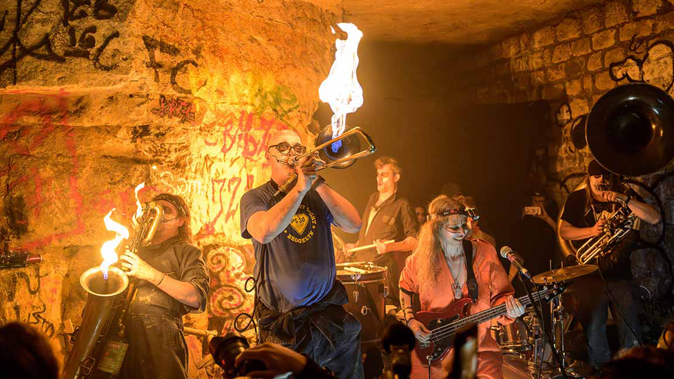 Band in graffitied cavern playing brass instruments with big flames coming out.