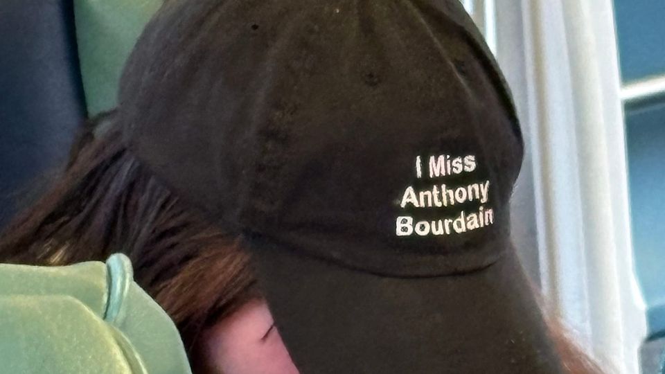 Person with head tilted down so you can't see their face. They are wearing ballcap reading "I Miss Anthony Bourdain"
