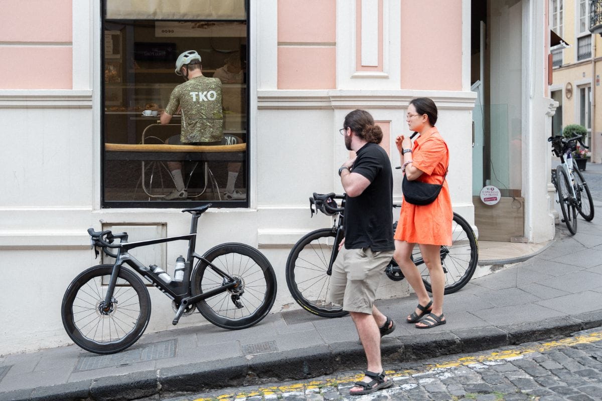 A cyclist seated in a café, with locals walking by in the foreground, showing the relaxed rural town vibe of Tenerife and its cycling culture.