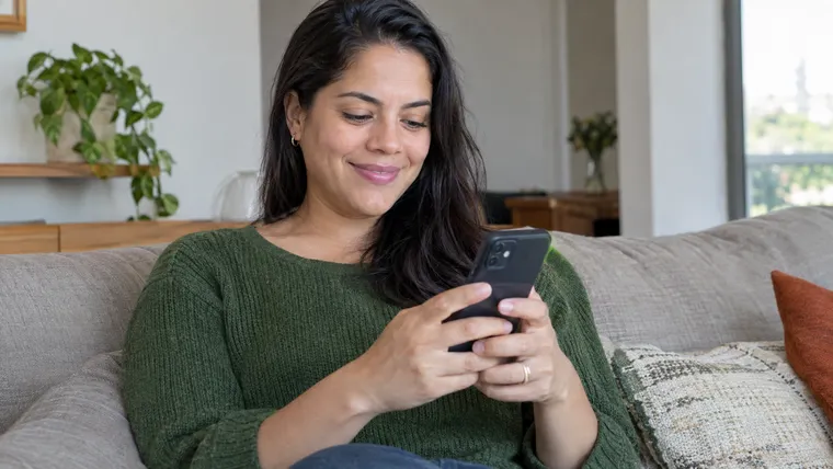 Imagem ilustrativa gerada por IA: mulher no sofá de casa usa o celular com expressão de alívio ao pedir o seguro-desemprego.