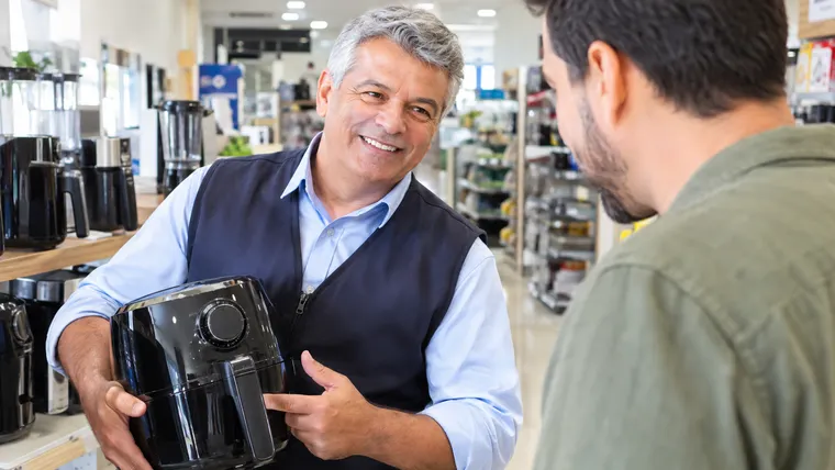 Imagem ilustrativa gerada por IA: vendedor de cabelos grisalhos atende cliente sorrindo em uma loja de eletrodomésticos.