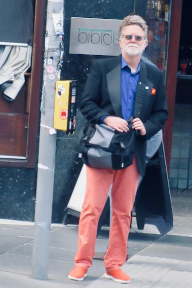 Image of a bearded man with grey hair, grey beard, and glasses. He is wearing orange shoes, orange trousers, a blue shirt, and a black tuxedo jacket while waiting at a street corner in Edinburgh Scotland, carrying a shoulder bag.