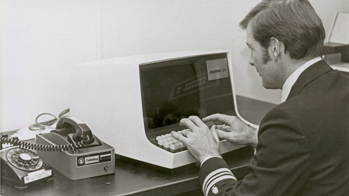 Black-and-white photo of man in a Naval uniform typing on a 1980’s style terminal, with a telephone in an acoustic modem next to him.