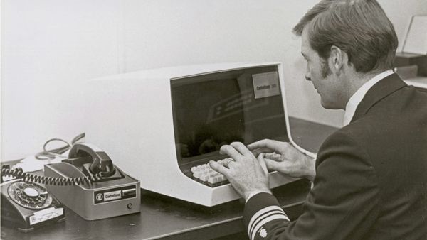 Black-and-white photo of man in a Naval uniform typing on a 1980’s style terminal, with a telephone in an acoustic modem next to him.