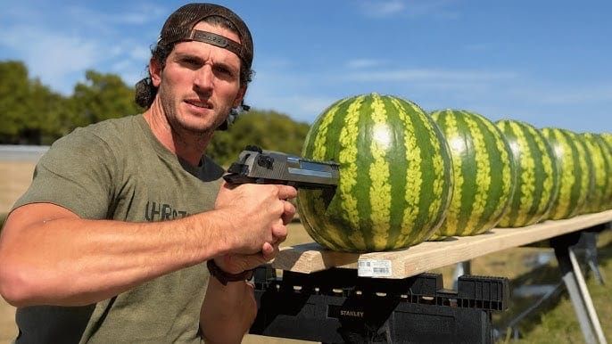 A man pointing a big gun at a line of watermelons