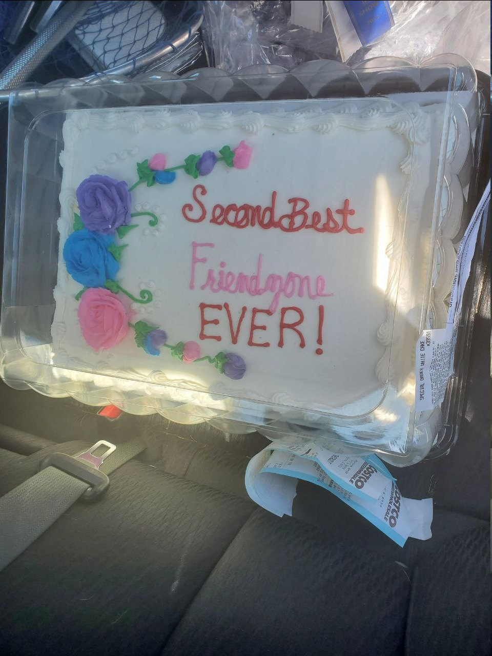 Close-up of a white sheet cake in a clear plastic clamshell, sitting on a car's back seat next to the seatbelt buckle, sunlight flaring across the lid. Icing script in red and pink reads "Second Best Friendzone EVER!" across three lines, beside a curve of purple, blue, and pink frosting roses. A label on the container reads "SPECIAL ORDER WHITE CAKE," and a Costco receipt is tucked underneath.