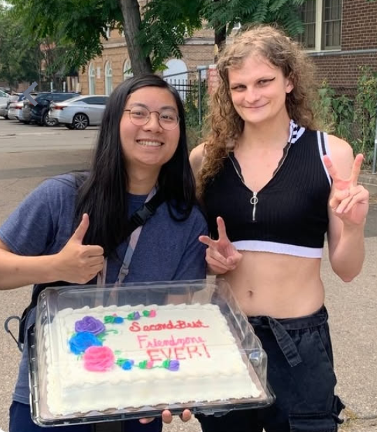 Me and Lux standing outside on a sunny street, holding a white sheet cake in a clear plastic box. Pink cursive icing on the cake reads "Second Best Friendzone EVER!", next to purple and pink frosting roses. I'm holding the cake and giving a thumbs up; Lux is throwing double peace signs. We're both grinning.
