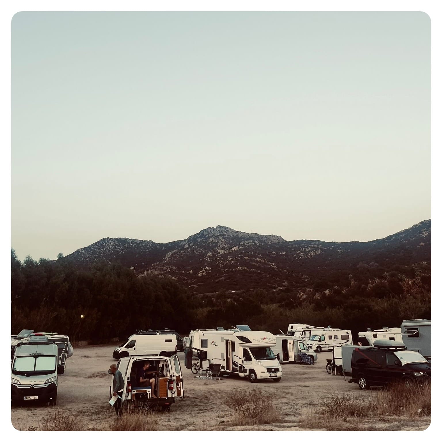 van life cluster of vans and motorhomes and RVs parked on a dirt parking lot next to beach