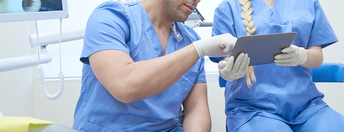 Two dental professionals in blue scrubs reviewing a tablet together in a clinic.