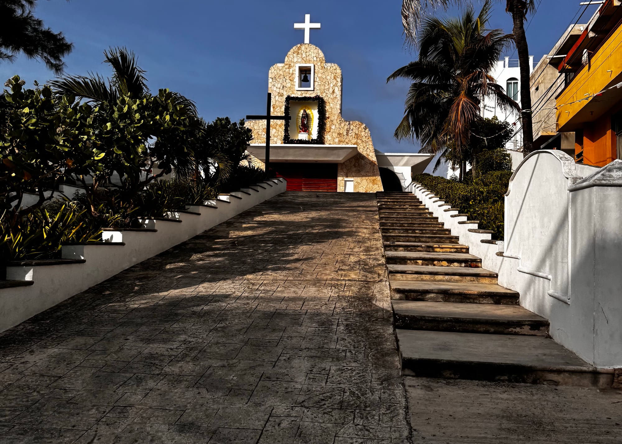 The Chapel of Guadalupe sits atop a small hill in Isla Mujeres beneath dramatic storm clouds, surrounded by palm trees and tropical landscaping