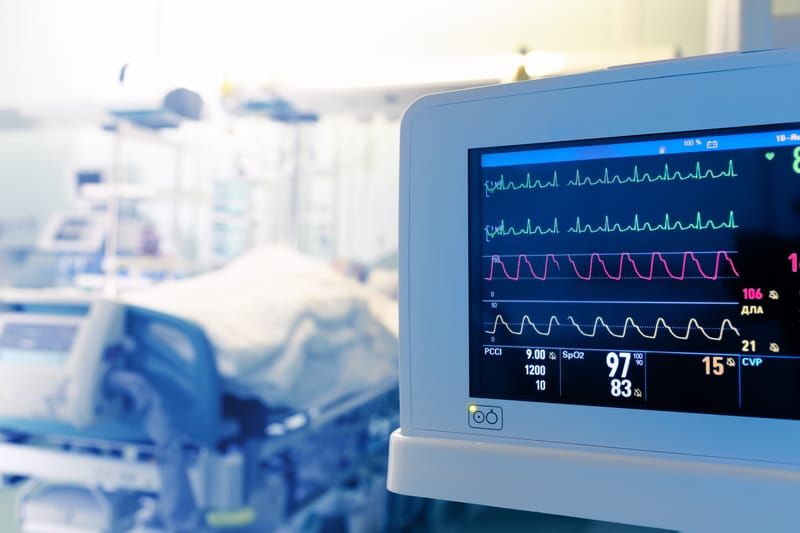 A hospital intensive care unit with a patient bed blurred in the background and a bedside monitor displaying heart rhythm, oxygen saturation, and vital signs in the foreground.