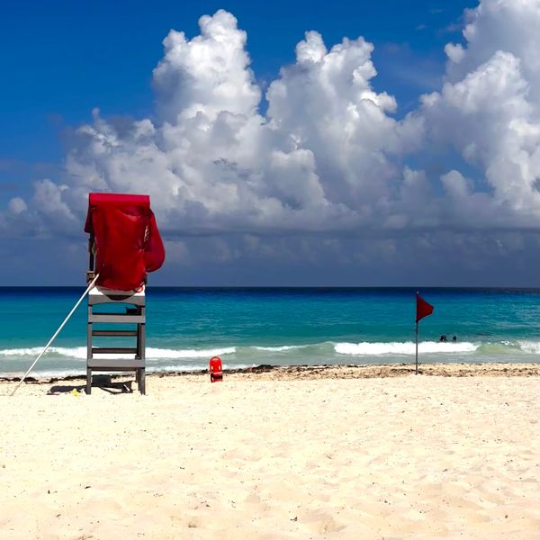 A white sand beach in Cancún with a lifeguard chair draped in red fabric, turquoise water, a red beach safety flag, and towering white clouds against a bright blue sky.