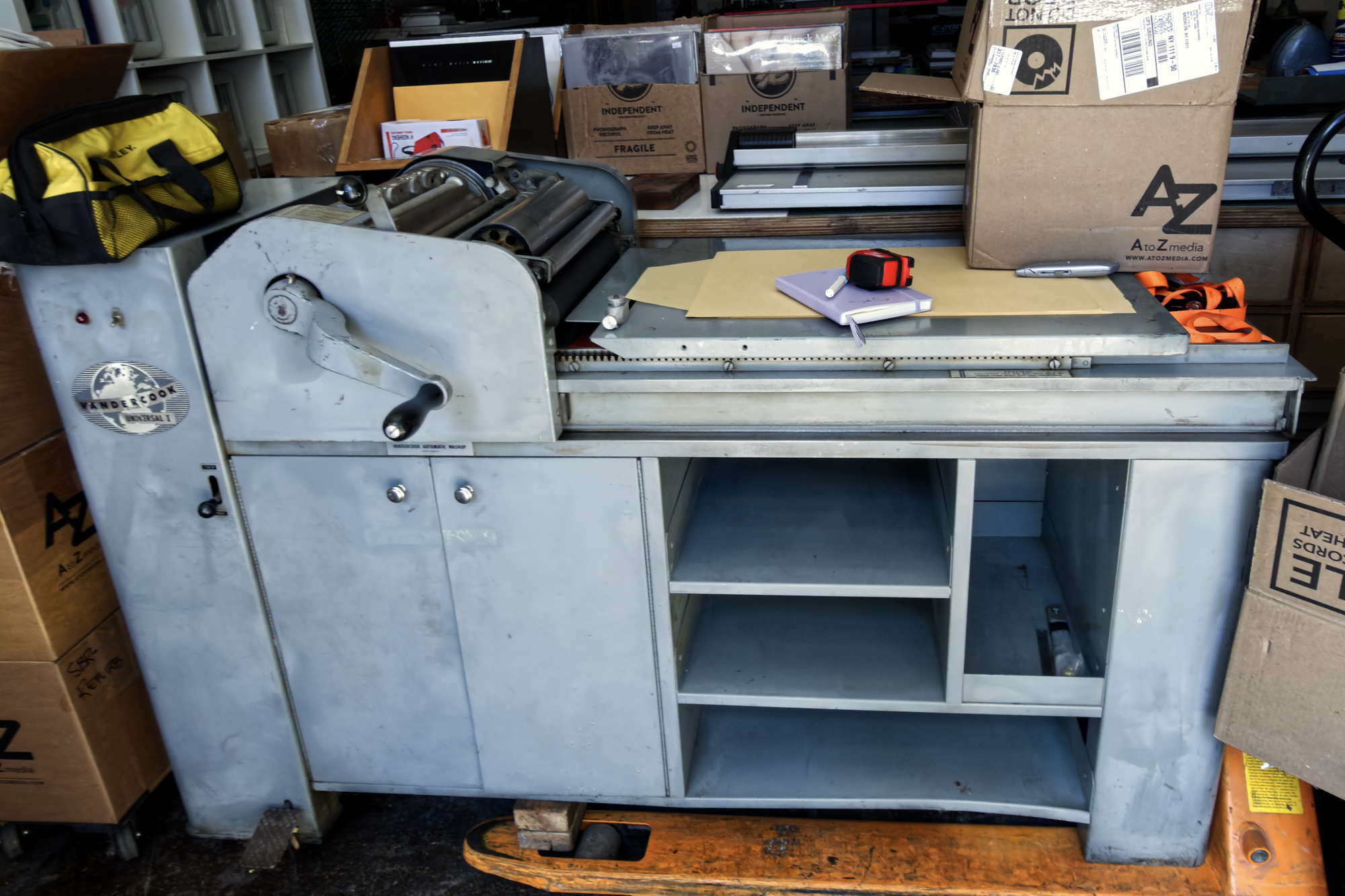 A vandercook universal 1 sits atop a bright orange palette joke supported by one leg and two two by fours. The other side of the press dangles off the edge