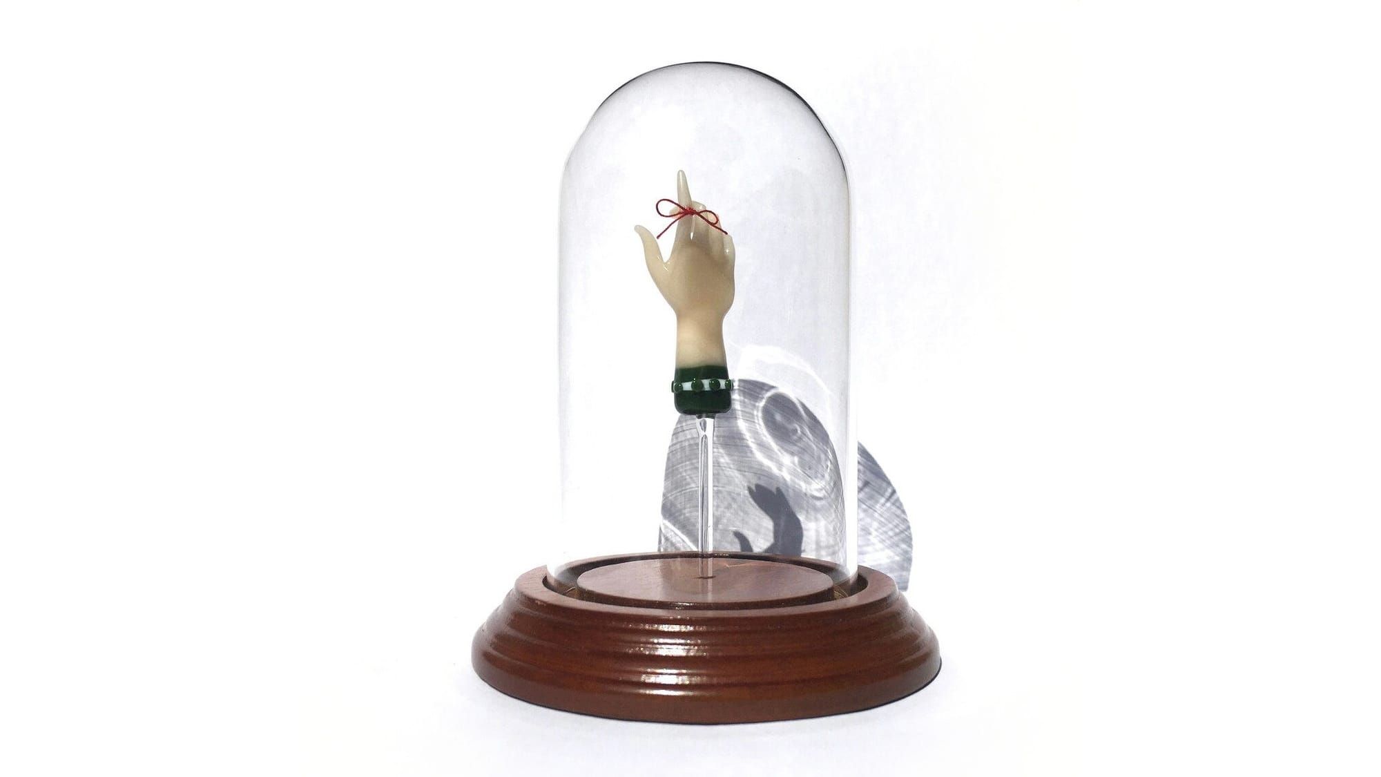 A hand with a green sleeve reaches made of borosilicate glass reaches upward with the index finger extended. A dainty red ribbon is tied around the index finger. Mounted in a glass cloche