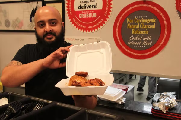 A guy with a big beard holding a to-co container filled with cooked chicken pieces.