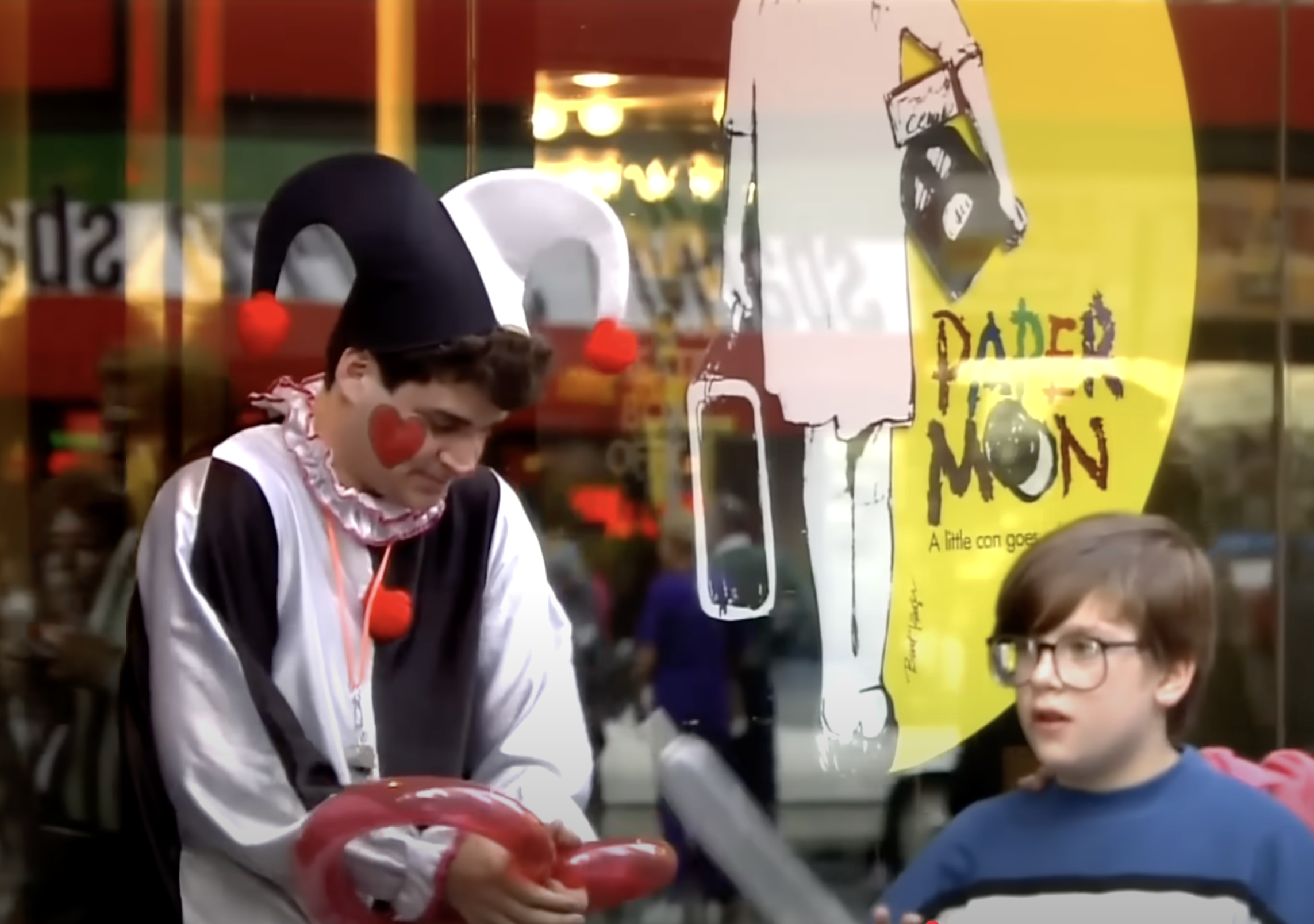 A kid in a jester costume making a balloon animal (I think it's a... circle) across the street from a Sbarro's.