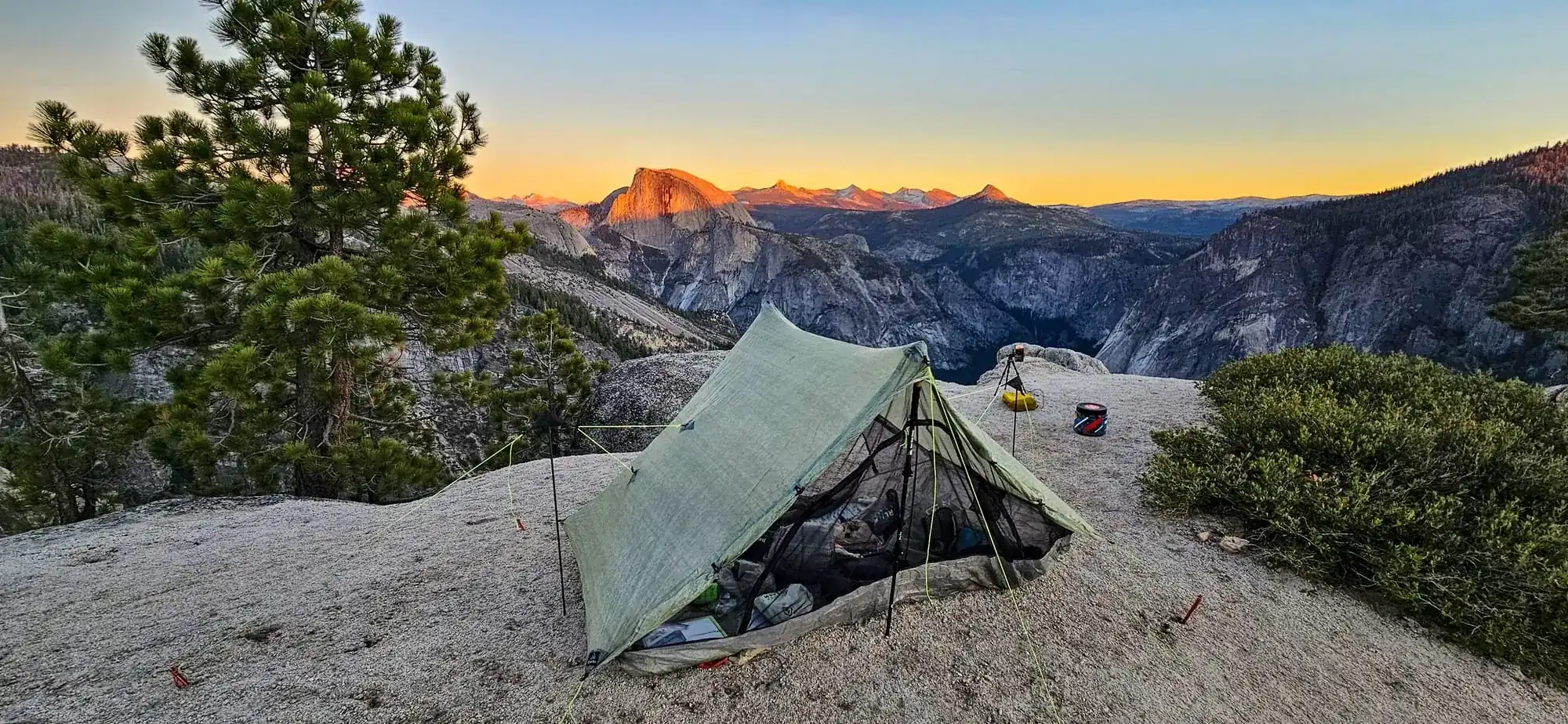 Backpacking camp with tarp tent setup on granite ledge overlooking Half Dome at sunset in Yosemite wilderness