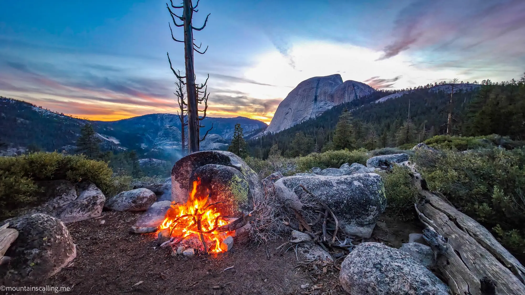 Campfire at dusk with granite dome and mountains visible during Clouds Rest backpacking trip in Yosemite wilderness