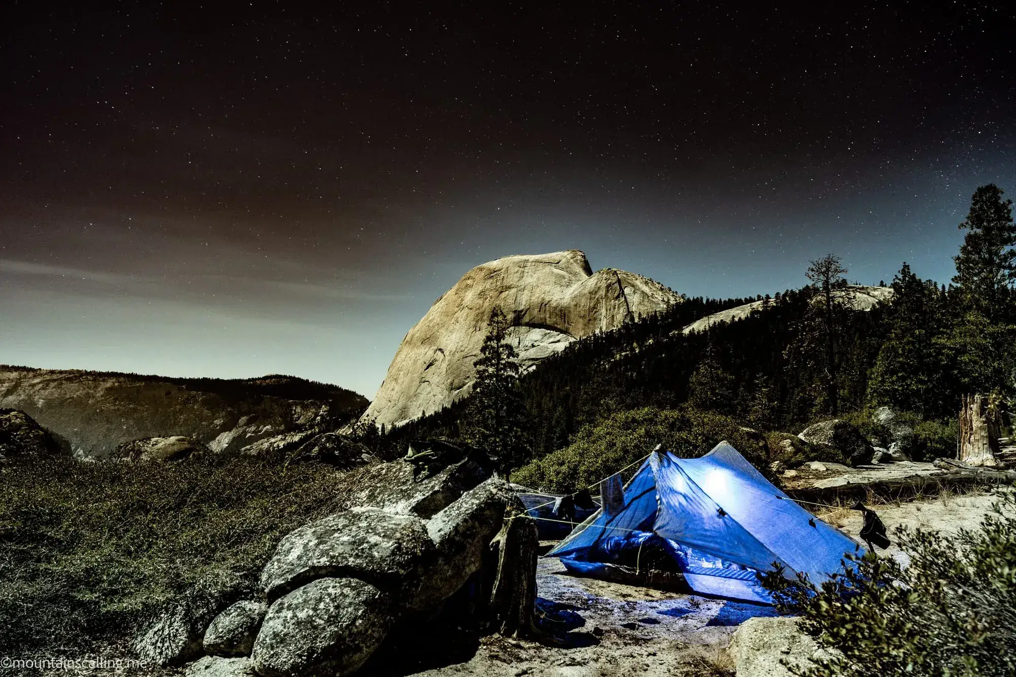 Blue tent under starry night sky with Half Dome granite formation illuminated in background at Yosemite backpacking camp