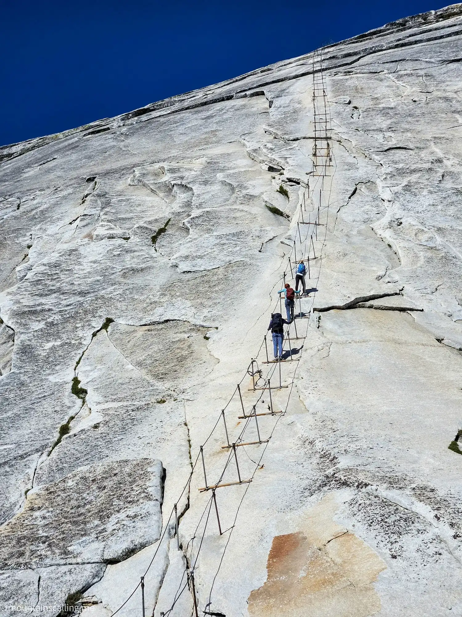 Hikers climbing Half Dome cables section with metal ladder system on steep granite face in Yosemite